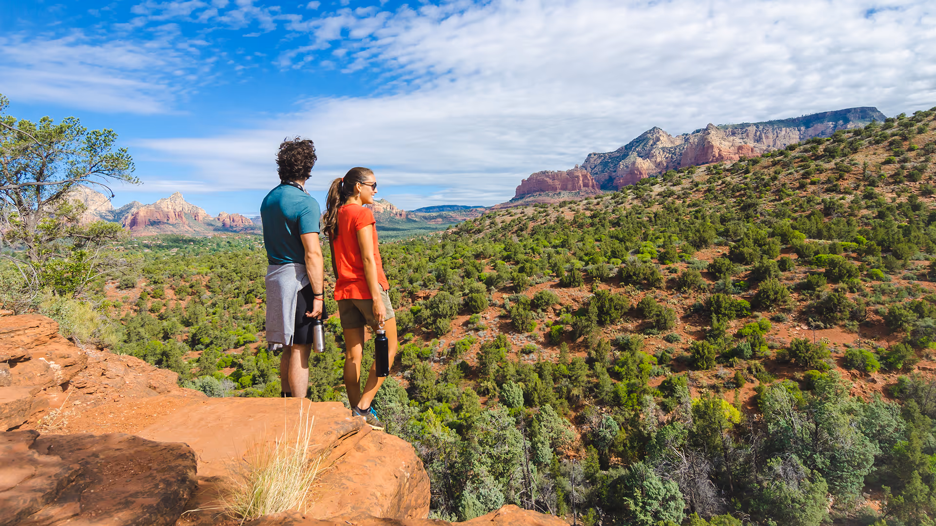 Couple with water bottles standing on a red rock cliff overlooking a green forest and red rock mountains under a partly cloudy sky.